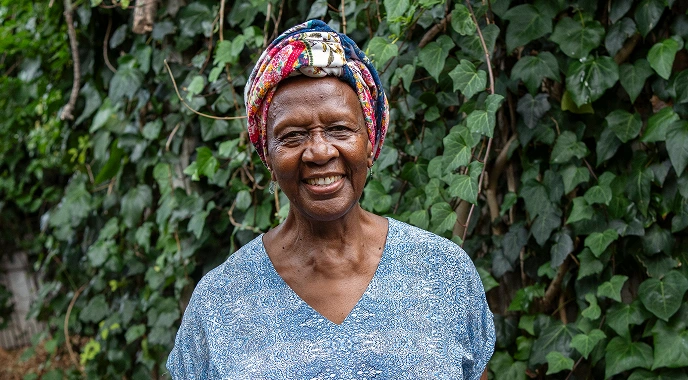 An elderly woman with a colorful headscarf smiles warmly, standing in front of lush greenery, representing joy and resilience.